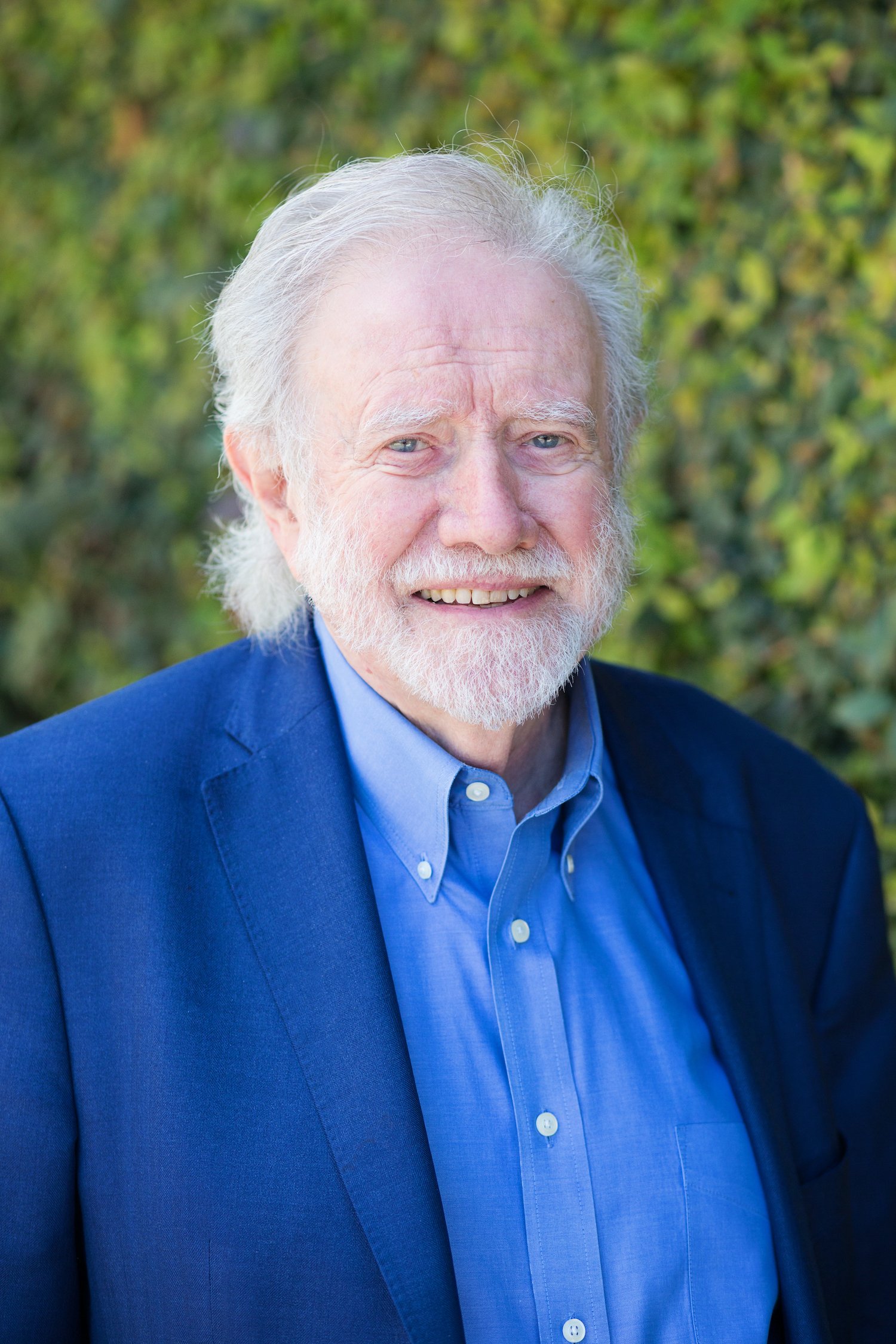 Smiling older man with beard in a blue suit and shirt standing outdoors with a leafy background.