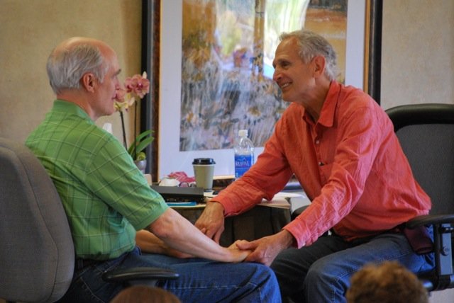 Two men sitting face to face in a warm indoor setting, holding hands and smiling at each other, with a small table, flowers, and a large window in the background.