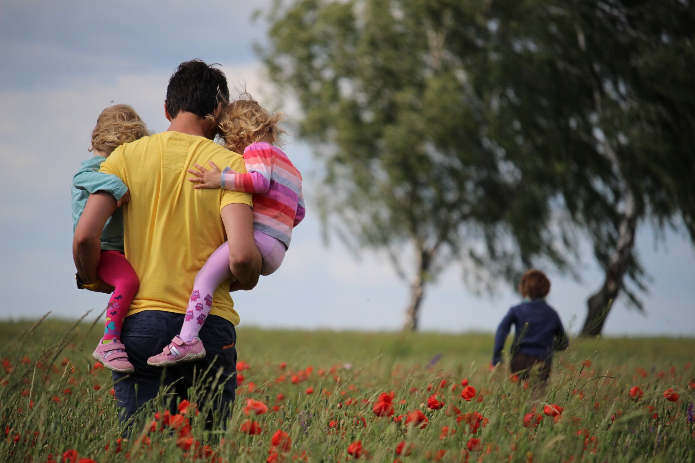 A man carrying two young children on his shoulders through a field of red flowers with a boy walking ahead near trees under a cloudy sky.