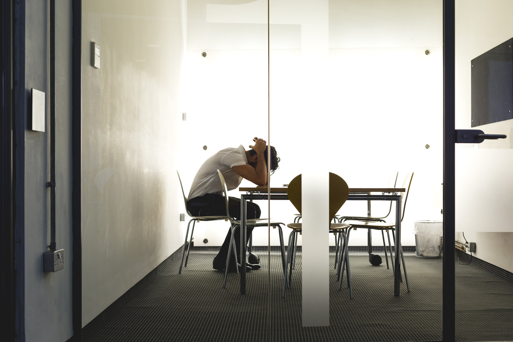 A person sitting at a conference table in a room with white walls and a large backlit window, leaning forward with head resting on hands.