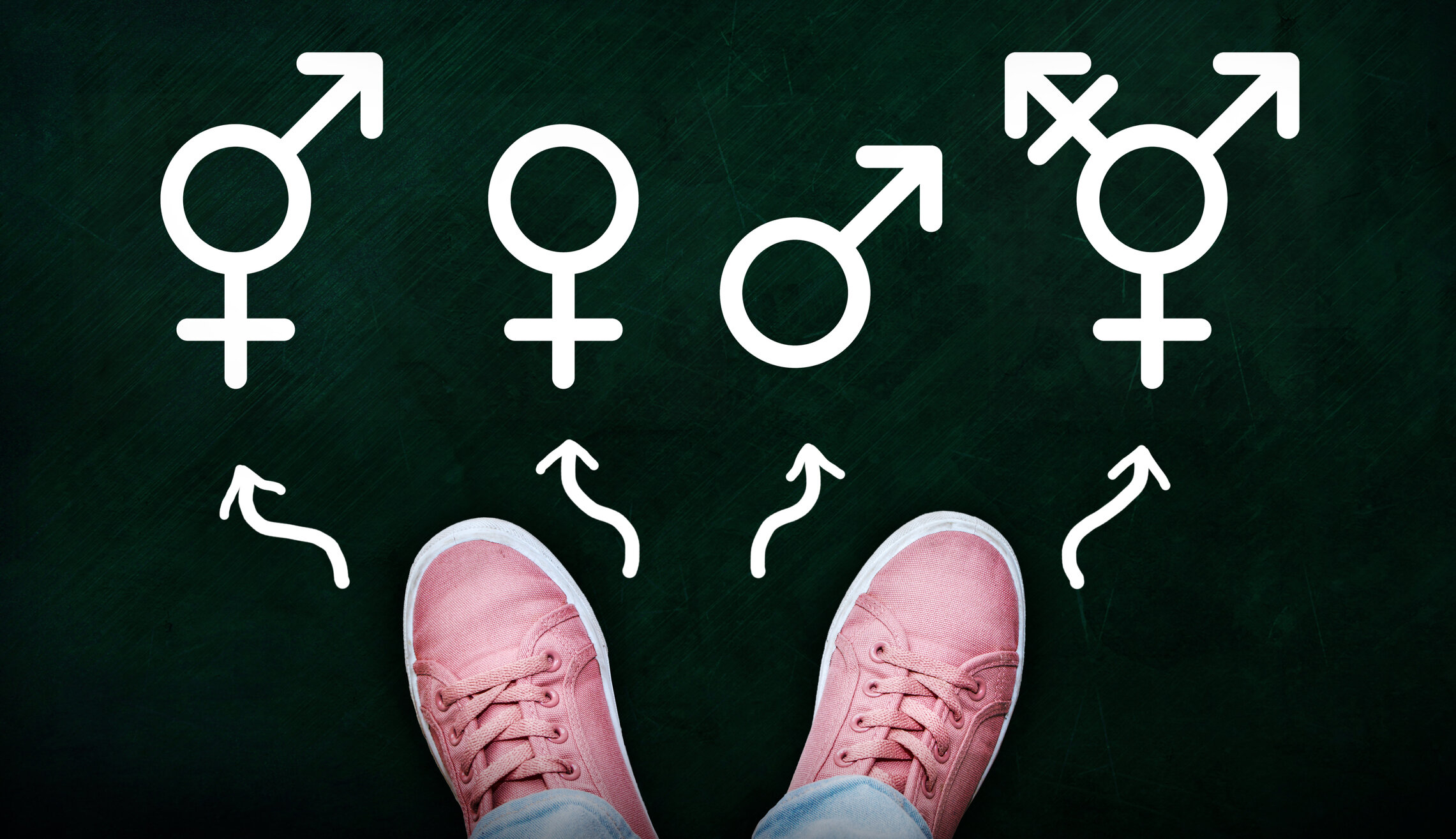 Top-down view of a person wearing pink sneakers and blue jeans, standing on a green chalkboard with various gender symbols and arrows drawn on it.