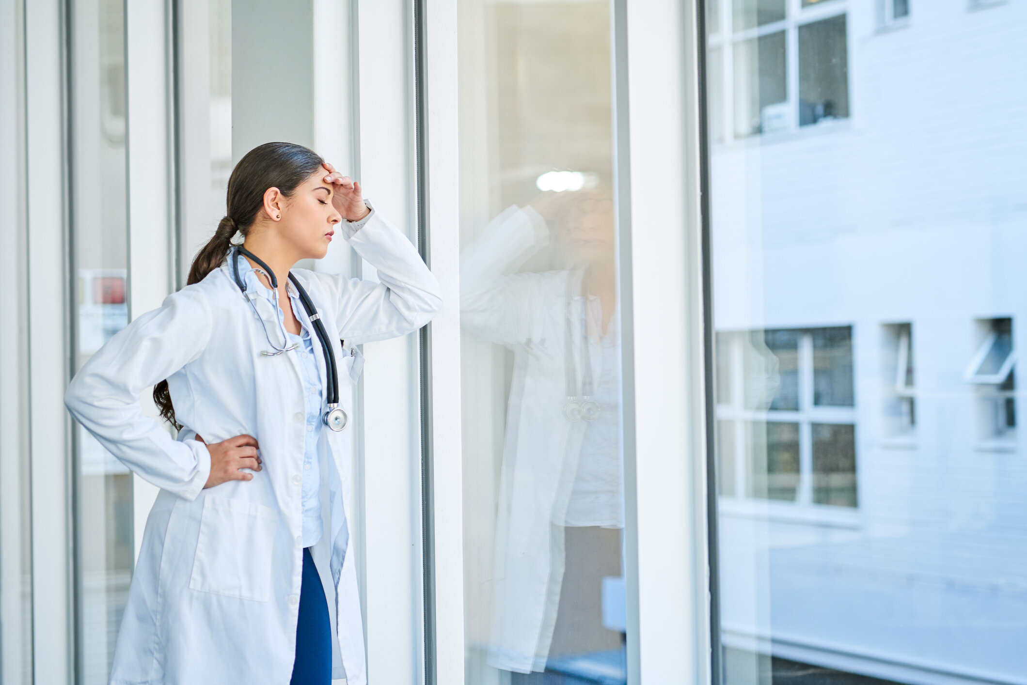 Female doctor or nurse in white coat with stethoscope shows a sign of headache or stress by holding her head, standing outside a glass window.