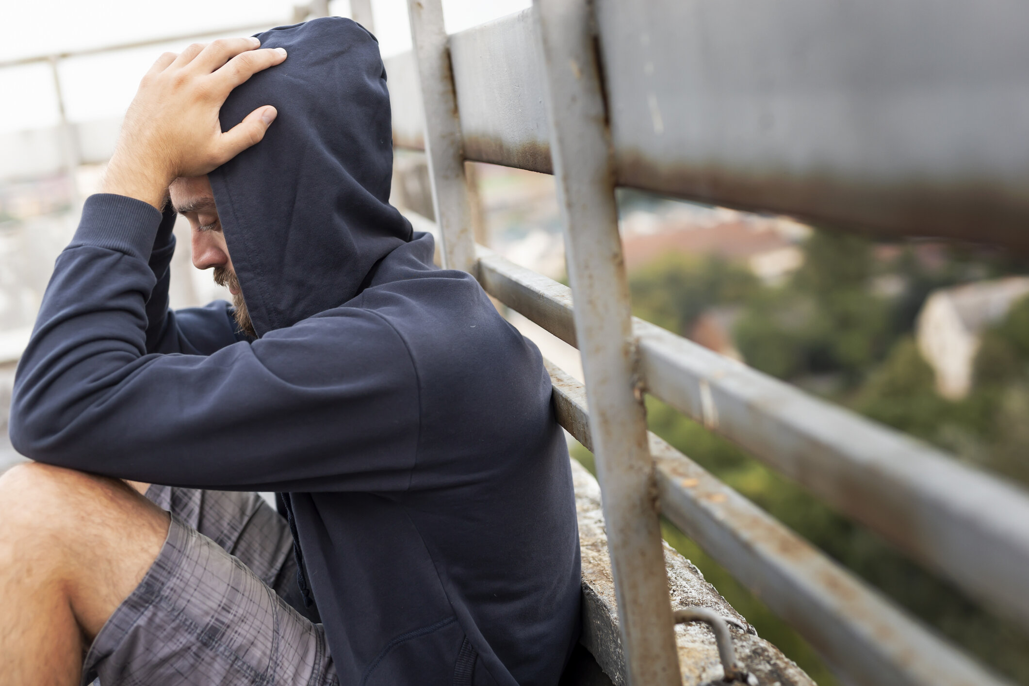Man sitting on a rooftop with a distressed expression, wearing a hoodie and shorts, holding his head in his hands.