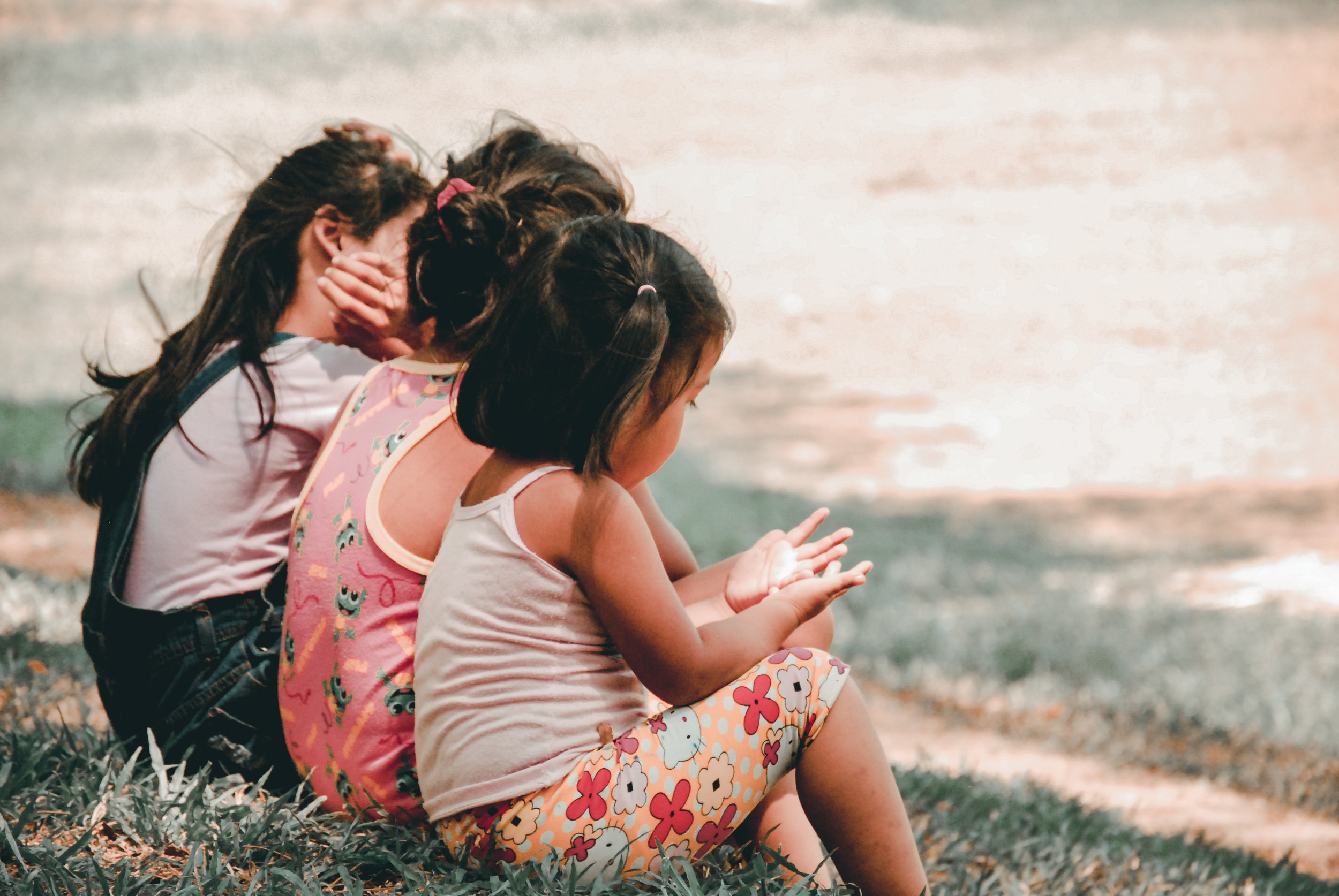 Four children sitting on grass near a water body, with three of them covering their ears with their hands and the fourth clapping.