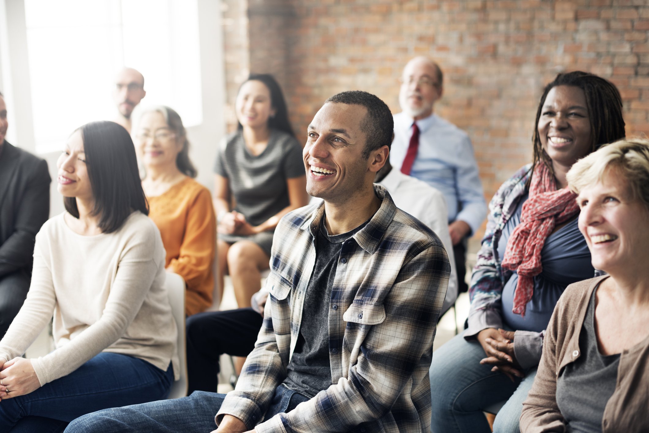 A diverse group of people sitting and smiling during a presentation or seminar in a bright room with brick walls.
