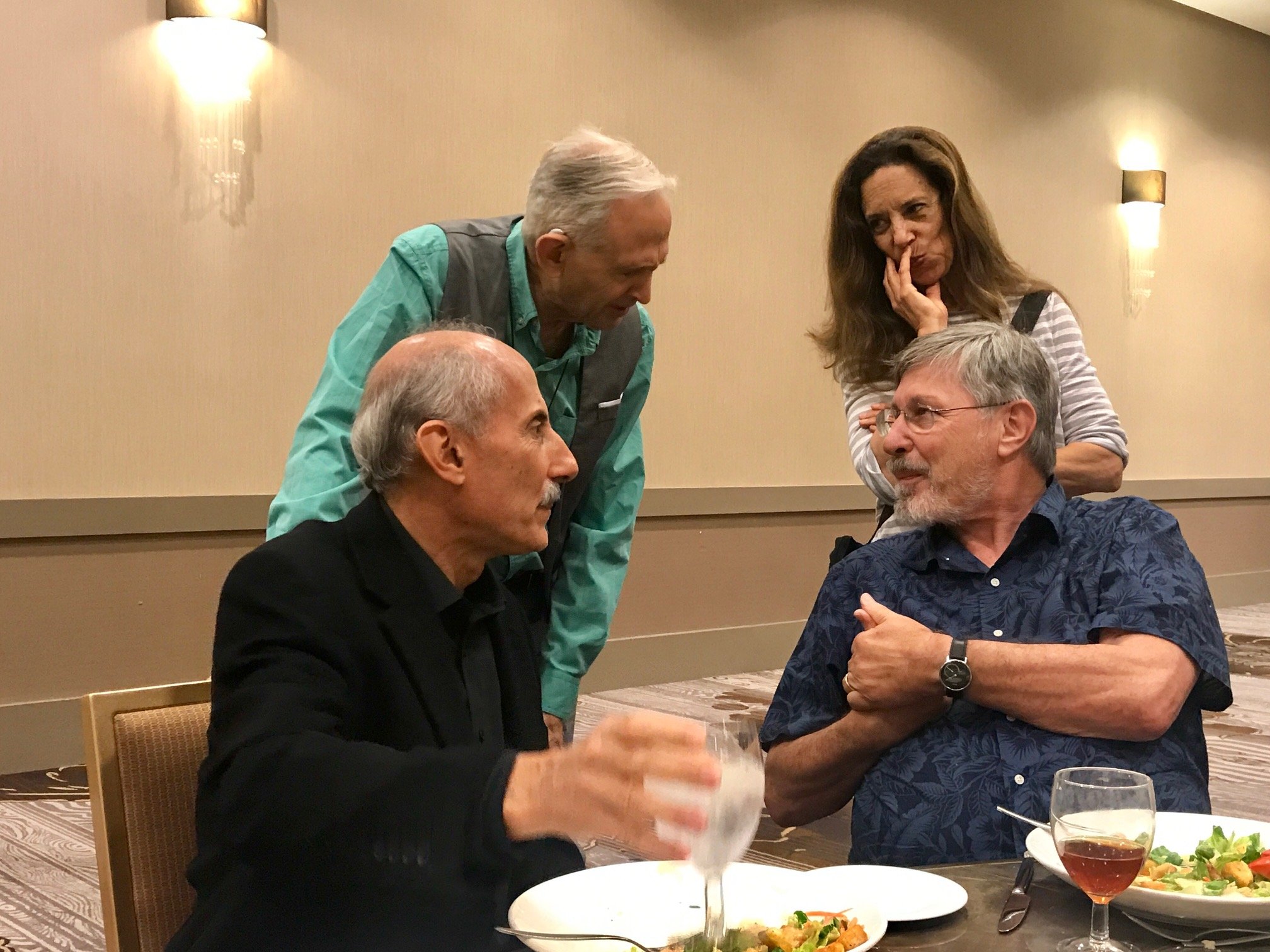 Jack Kornfield, Peter Levine, Bessel VanDerKolk  and Laura Regalbuto engaged in conversation at a dining table, with plates of food and drinks, in a banquet room with beige walls and wall-mounted lights.