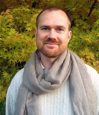 Man with a beard wearing a light-colored sweater and scarf standing in front of green and yellow foliage.