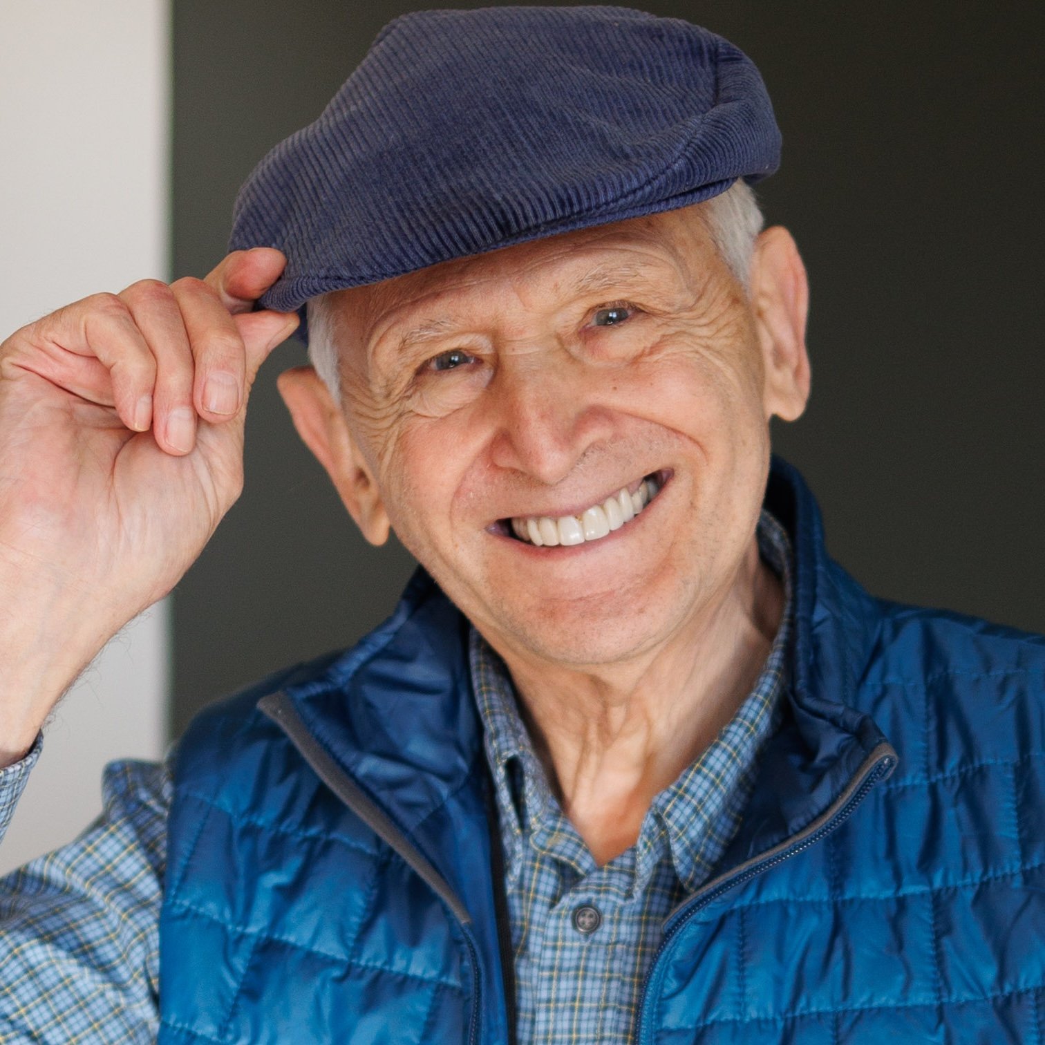 A smiling elderly man wearing a blue plaid shirt, a blue puffer vest, and a blue cap, holding the cap with his right hand.