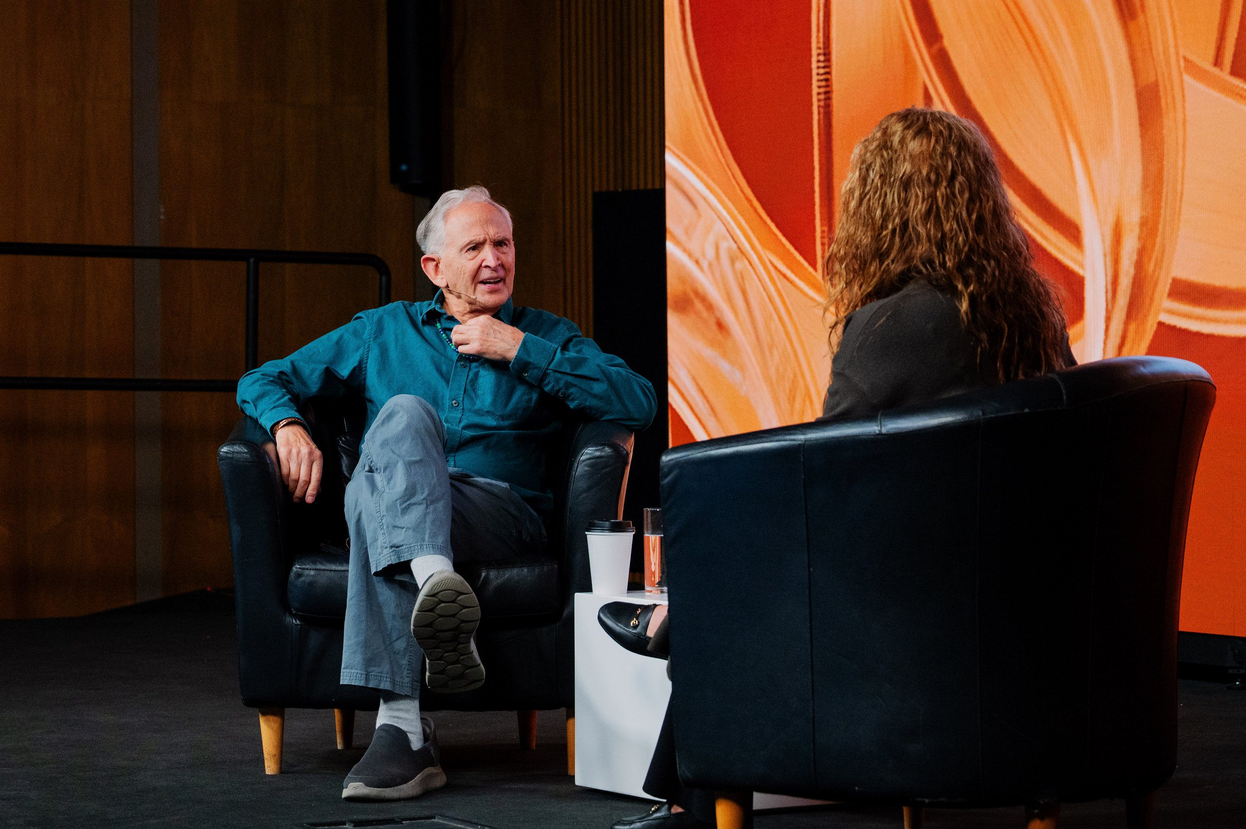 Dr Peter Levine wearing a teal shirt and gray pants sitting in a black armchair, speaking to a woman who is sitting in a black armchair in front of him. They are engaged in a conversation on a stage with a large orange and red background.