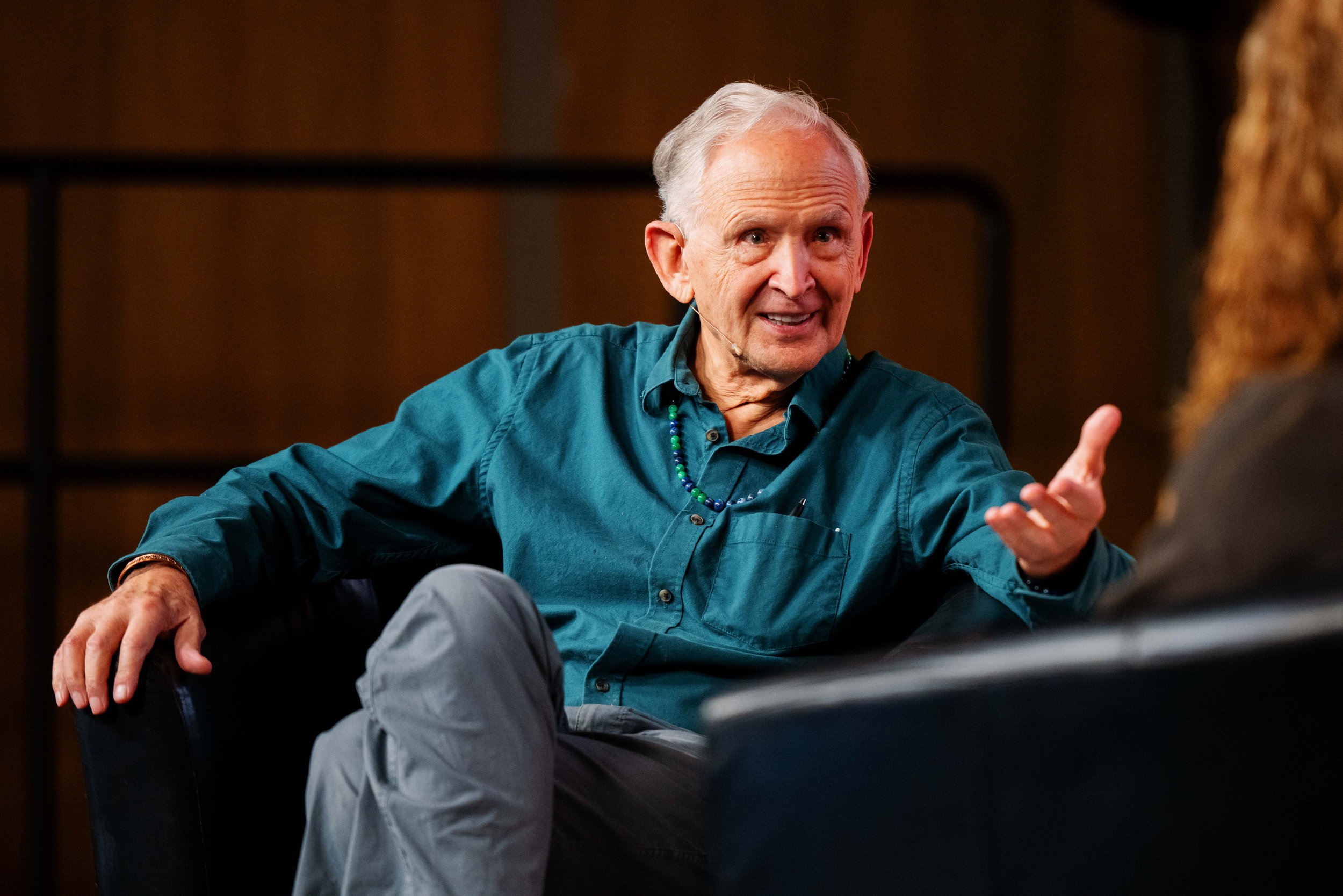 Dr Peter Levine wearing a teal shirt and gray pants, gesturing with his right hand during a conversation in a dimly lit indoor setting.