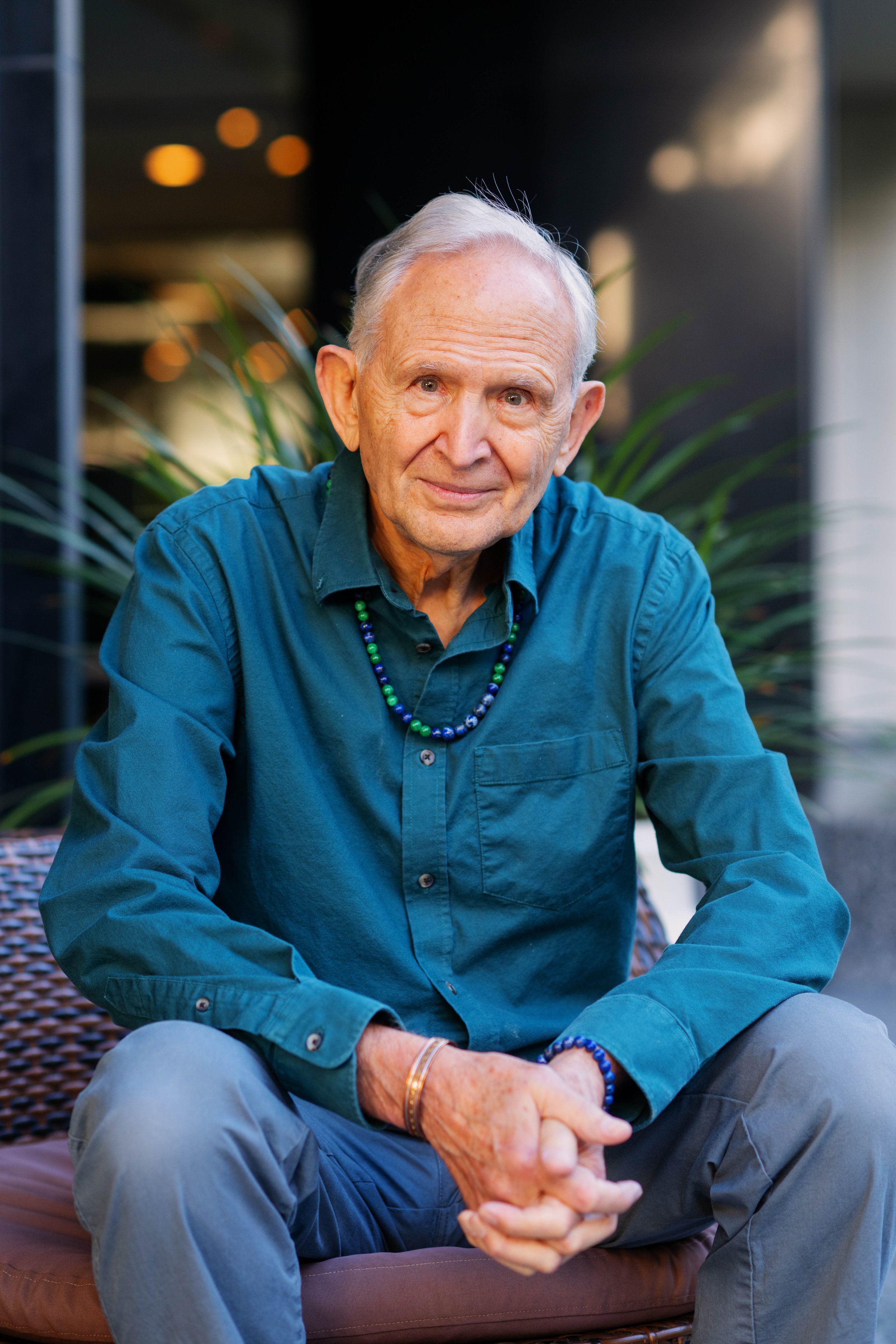 An elderly man with white hair and wearing a teal shirt, sitting outside on a wicker chair, smiling, with plants and lights in the background.