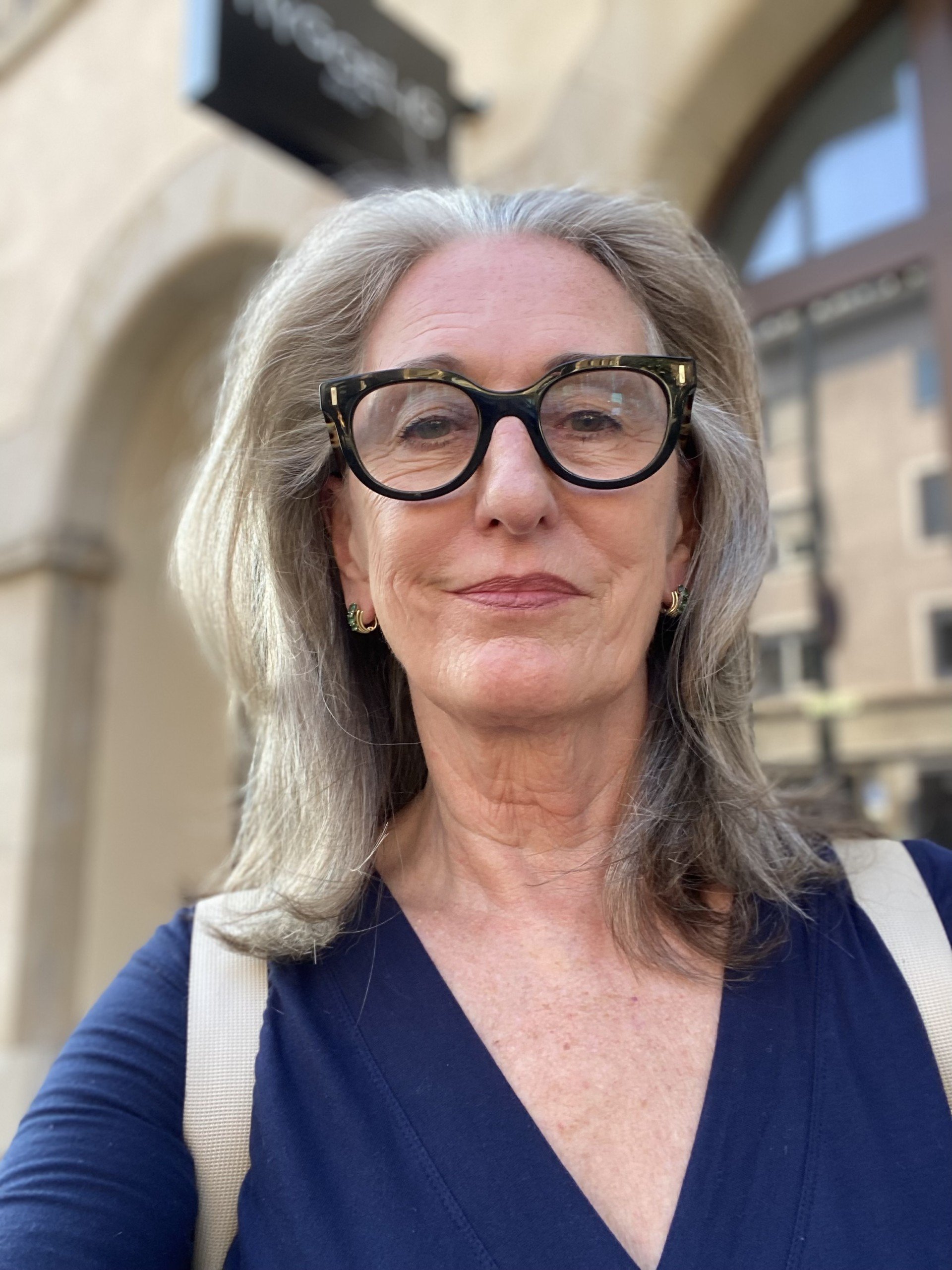 A woman with gray hair and black-rimmed glasses smiling outdoors in front of a blurred building background.